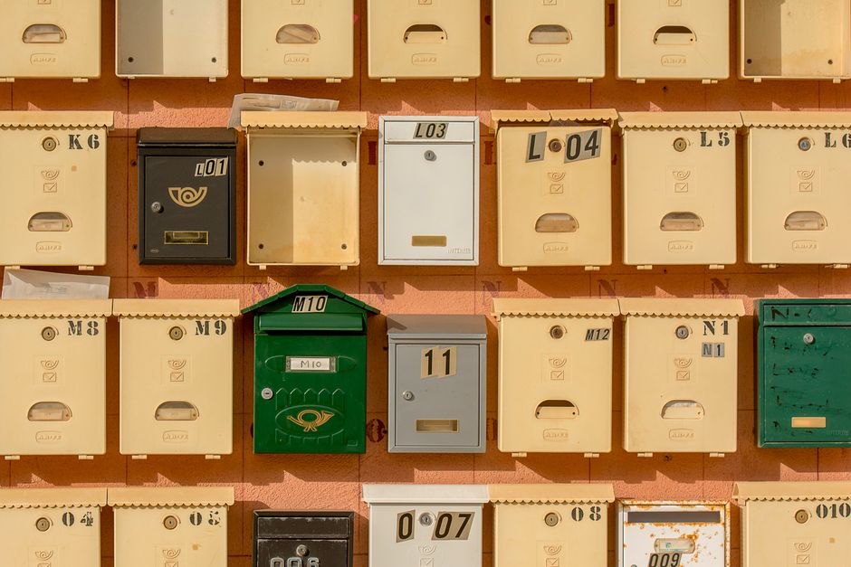 A close-up view of multiple postal mailboxes mounted on a wall, featuring various colors including beige, green, black, and gray. Each mailbox has a distinct letter or number label such as 'L01', 'L03