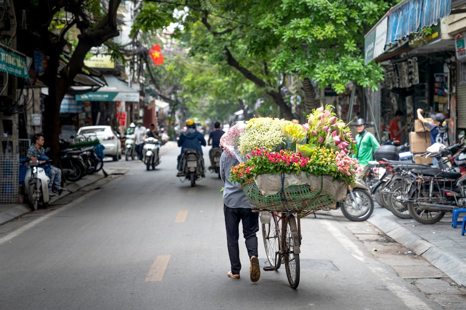 A street scene featuring a person pushing a bicycle cart filled with a large, colorful bouquet of fresh flowers, including pink lilies, white hydrangeas, and various green foliage. The arrangement is 