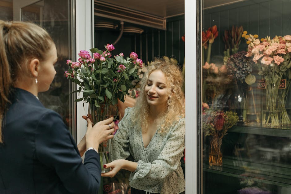 A woman with curly blonde hair wearing a light-colored blouse is receiving a bouquet of fresh pink roses from a florist at a flower shop. The florist, dressed in a dark blazer, is holding the floral a