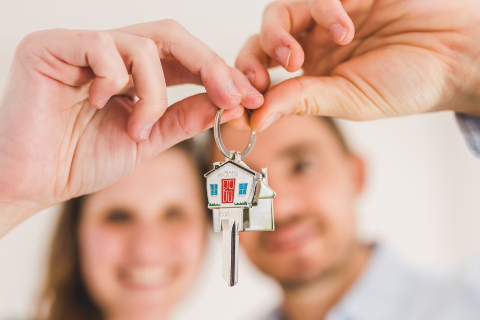 A close-up image of a person's hand holding a silver keyring with a decorative house-shaped keychain, symbolizing home ownership or a new home. The background features a smiling couple, slightly out o