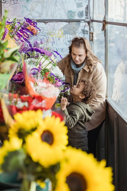 A woman and a young boy inside a flower shop, surrounded by a vibrant display of fresh floral arrangements. The woman has wavy brown hair and is wearing a beige coat over a blue turtleneck, while the