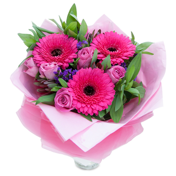 I see this bouquet as an expression of playful elegance: three vivid fuchsia Gerbera daisies open wide with confident, layered petals and dark centres that give the arrangement a bold focal point. Surrounding them are roses in a muted lavender-pink - the kind of dusty-rose that reads soft in daylight and feels almost powdery to the touch, their buds promising more bloom to come. Scattered amongst these are fine sprays of purple statice, lending a feathery, textural contrast that suggests wildflower edges drifting through a cultivated garden. The greenery beneath - broad, glossy leaves likely salal or eucalyptus - gives the composition a dense, verdant base and a faintly aromatic green note when handled. The bouquet is folded into a pale pink matte tissue inner wrap and finished with an outer sheet of translucent fuchsia-pink cellophane that shimmers without overpowering; the folds are neat and considered, the stems tucked so the whole feels secure for delivery. At the lower rim you can glimpse a clear glass vase, implying the flowers are intended for immediate display on a mantel or dining table. Picture this brought to a neighbour on St Paul's Road or laid on a bench in De Beauvoir - it radiates warmth, gratitude and gentle celebration. As a local florist accustomed to Canonbury's small pleasures, I would recommend this for Mothers' Day, a birthday surprise, or a quiet anniversary; its scent is restrained yet inviting, and its palette is designed to make anyone feel cherished.